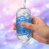 A hand holds the Tenga Spinner DX 01 Drops by Tenga, showcasing its blue spiral design as water is poured onto the transparent masturbator against a dreamy purple and pink blurred background.